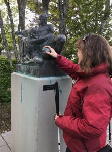 Photo of Amy standing in front of a bronze metal sculpture on a high cement pedestal. The sculpture is of two men wrestling, and Amy is touching a knee of one of the figures.
